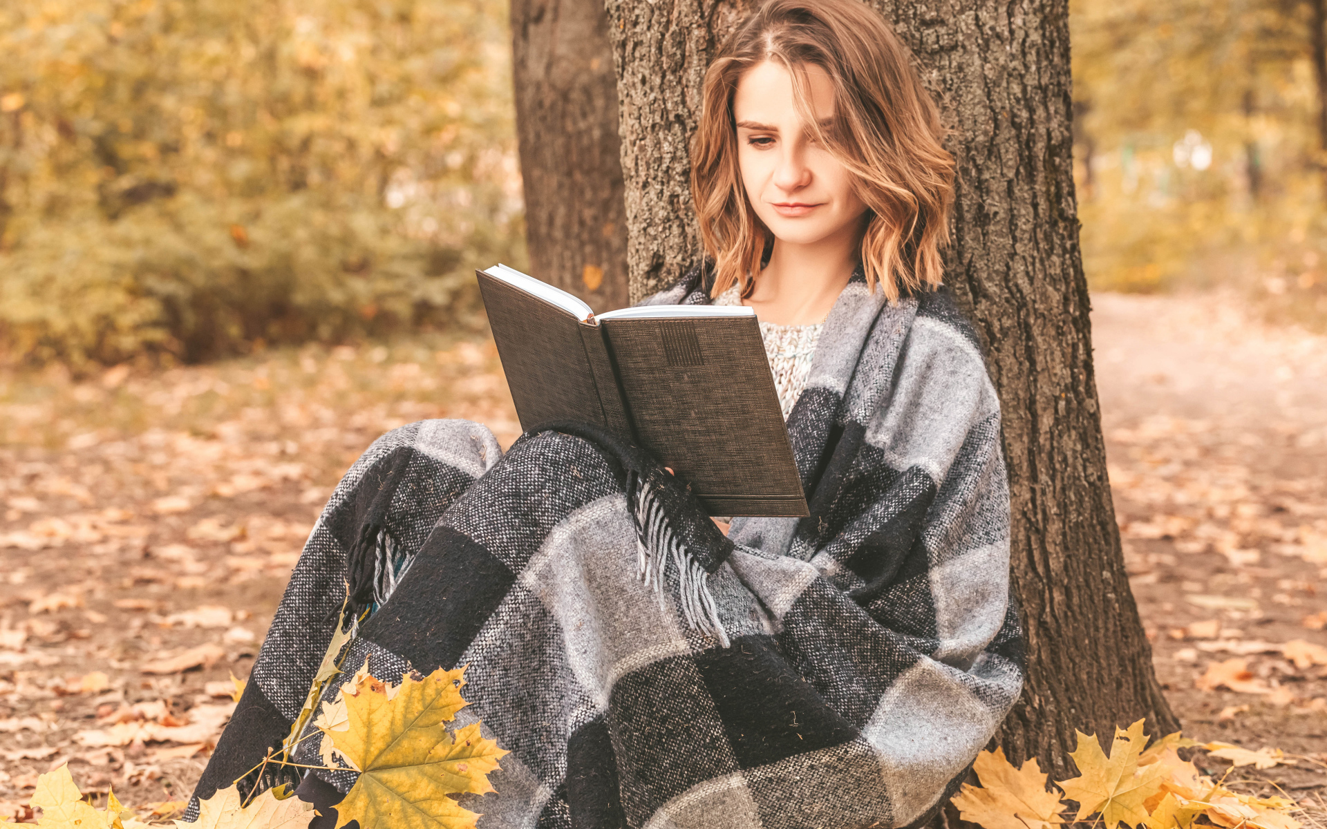 Девушка в пледе сидит с книгой под деревом