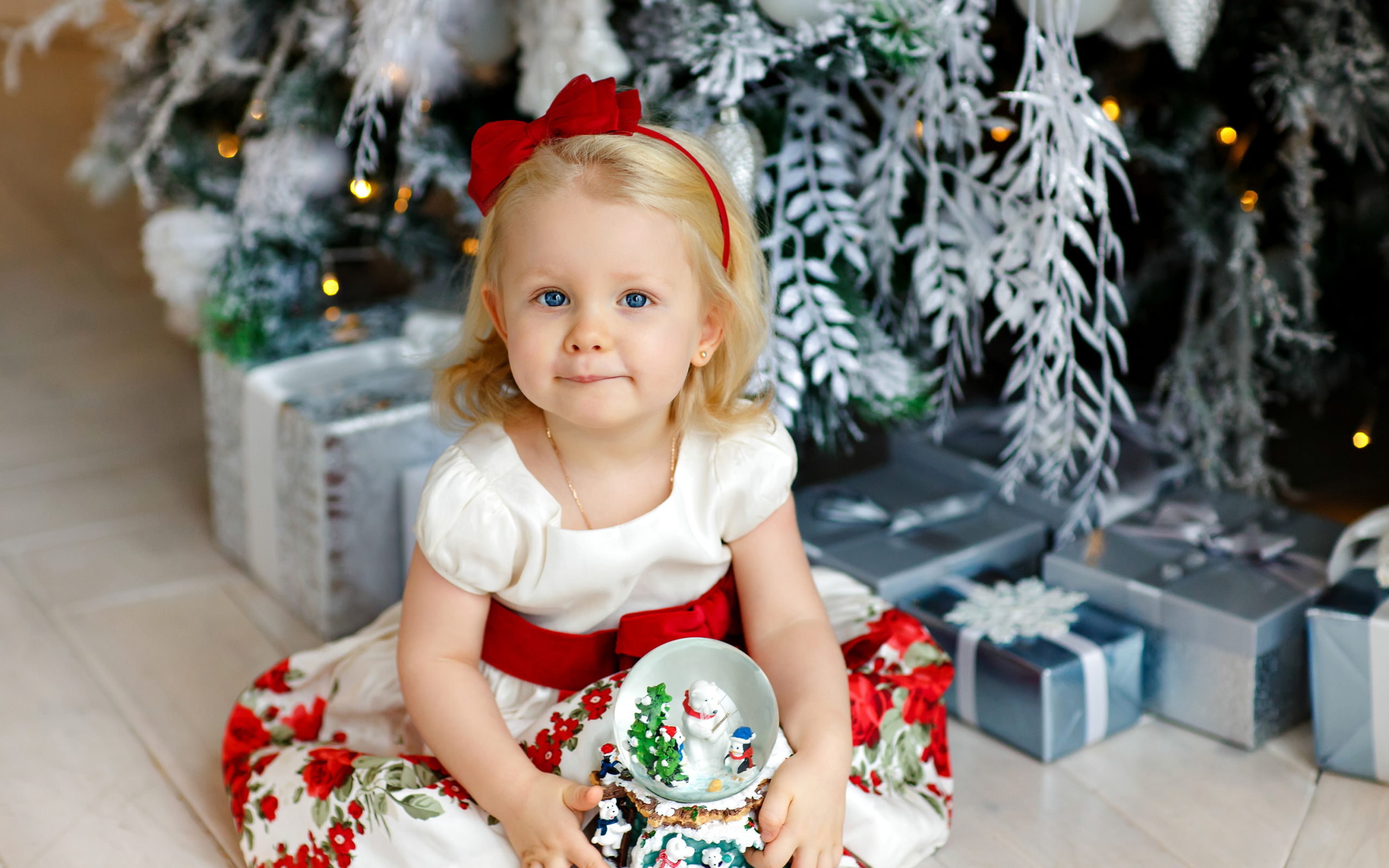 Маленькая девочка блондинка сидит с подарками под новогодней елкой