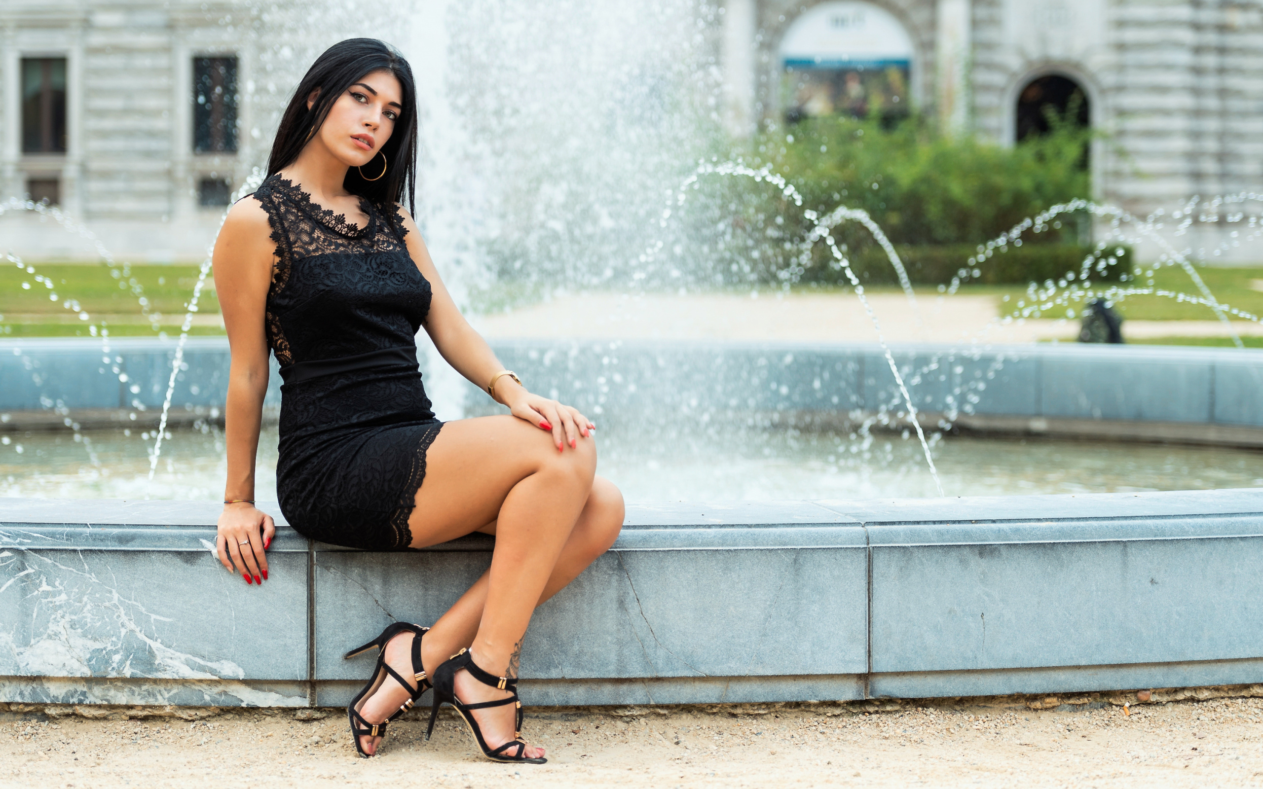 Красивая брюнетка в черном платье сидит у фонтана