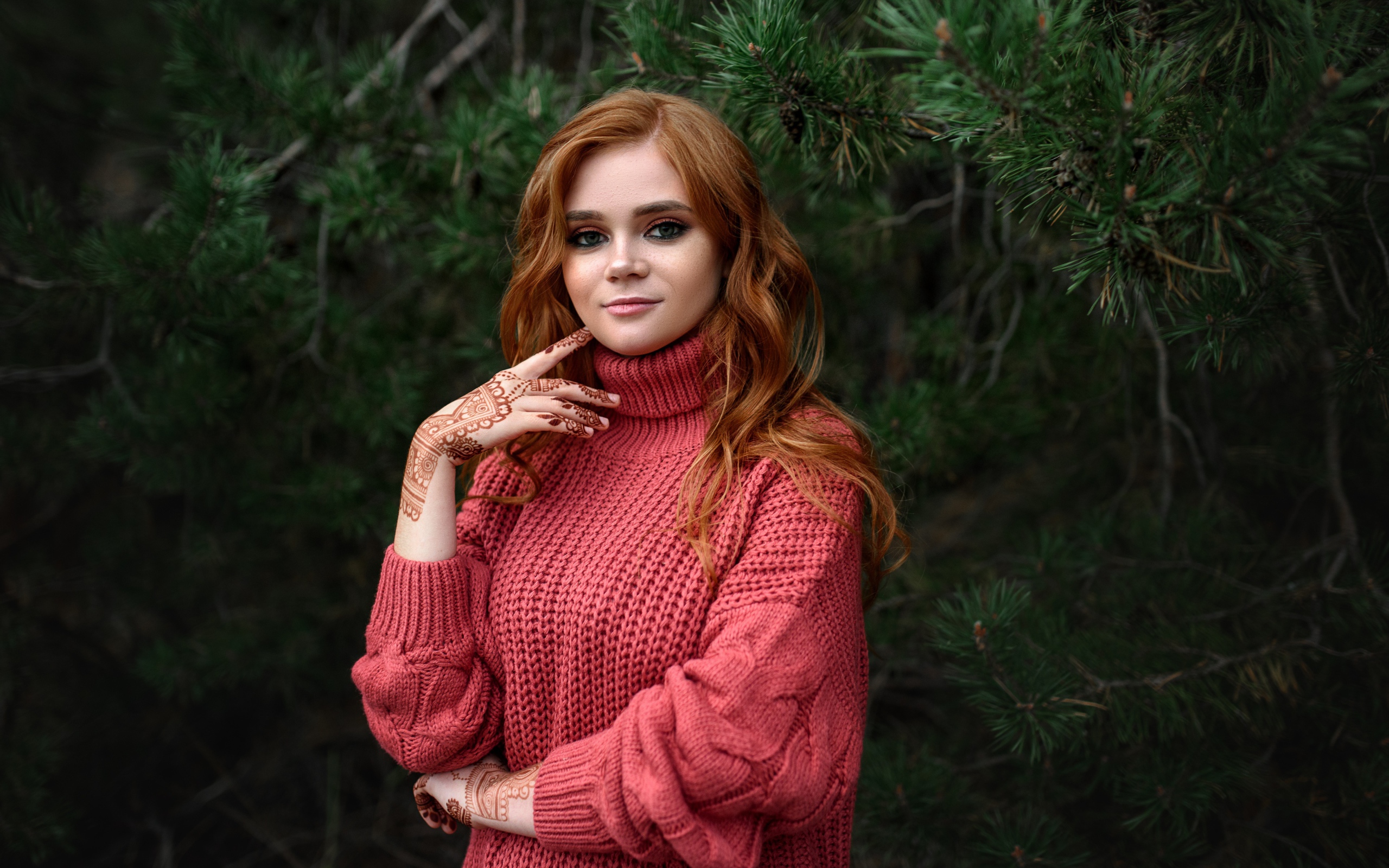 Рыжеволосая девушка в розовом свитере стоит у ели