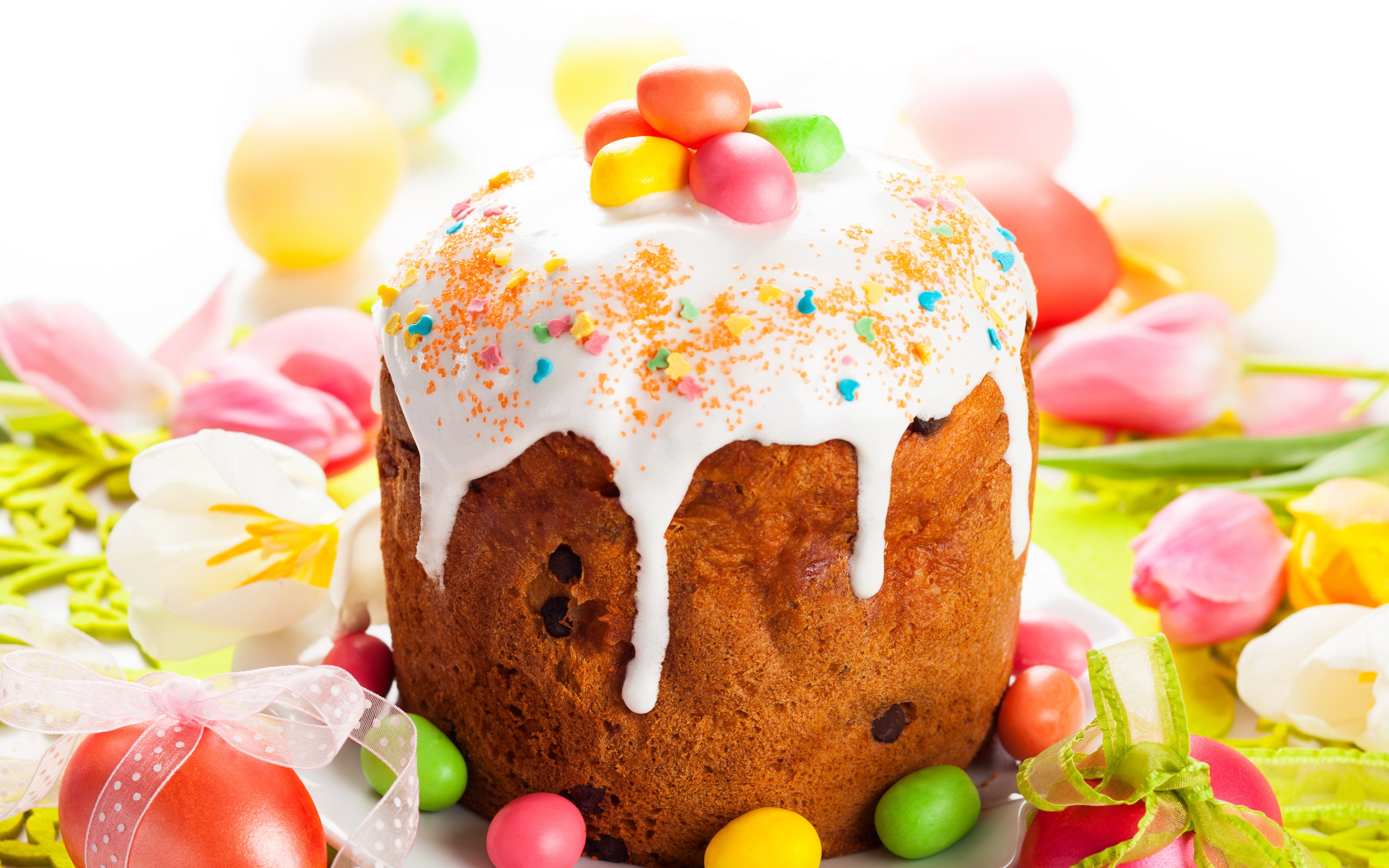 Красивый пасхальный кулич с белой глазурью и конфетами на столе с яйцами