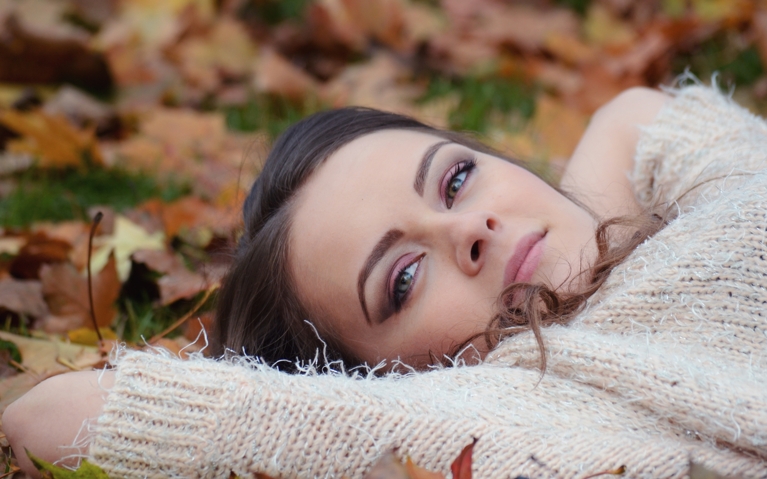 Красивая девушка в теплом свитере лежит на сухой листве 