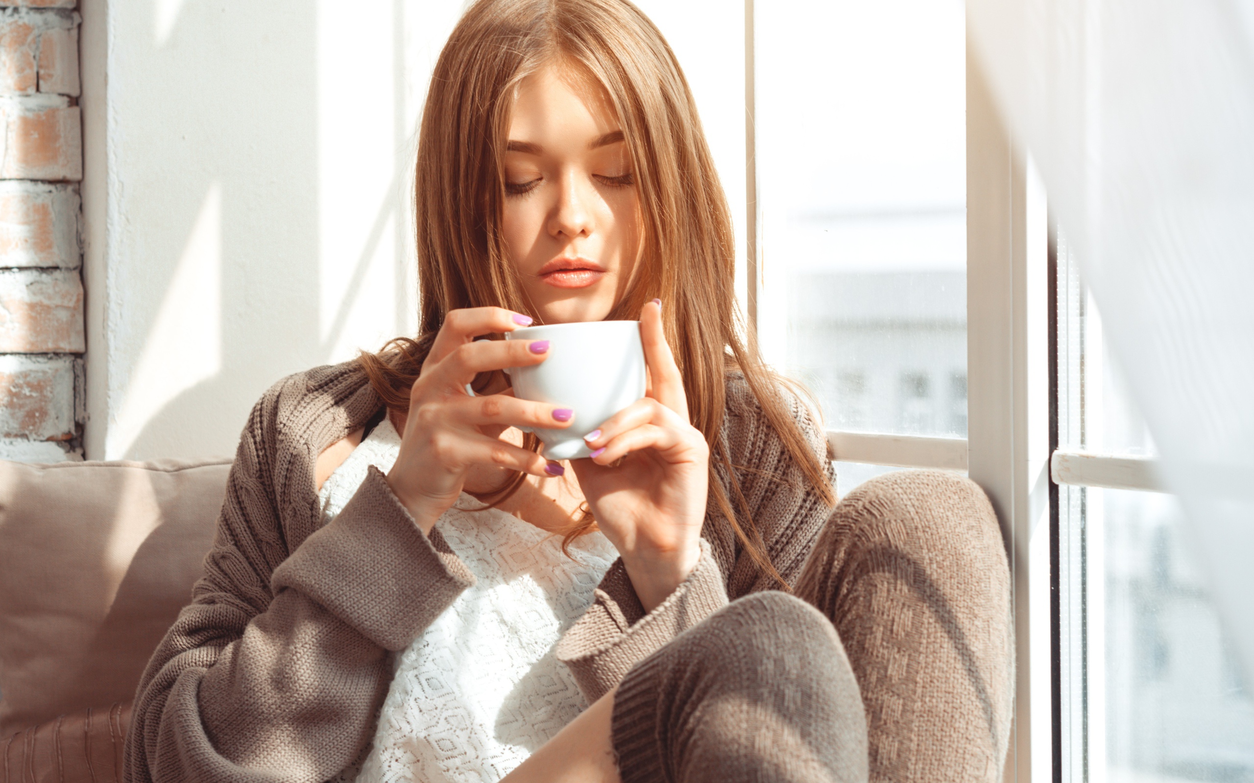 Красивая девушка в свитере пьет кофе у окна