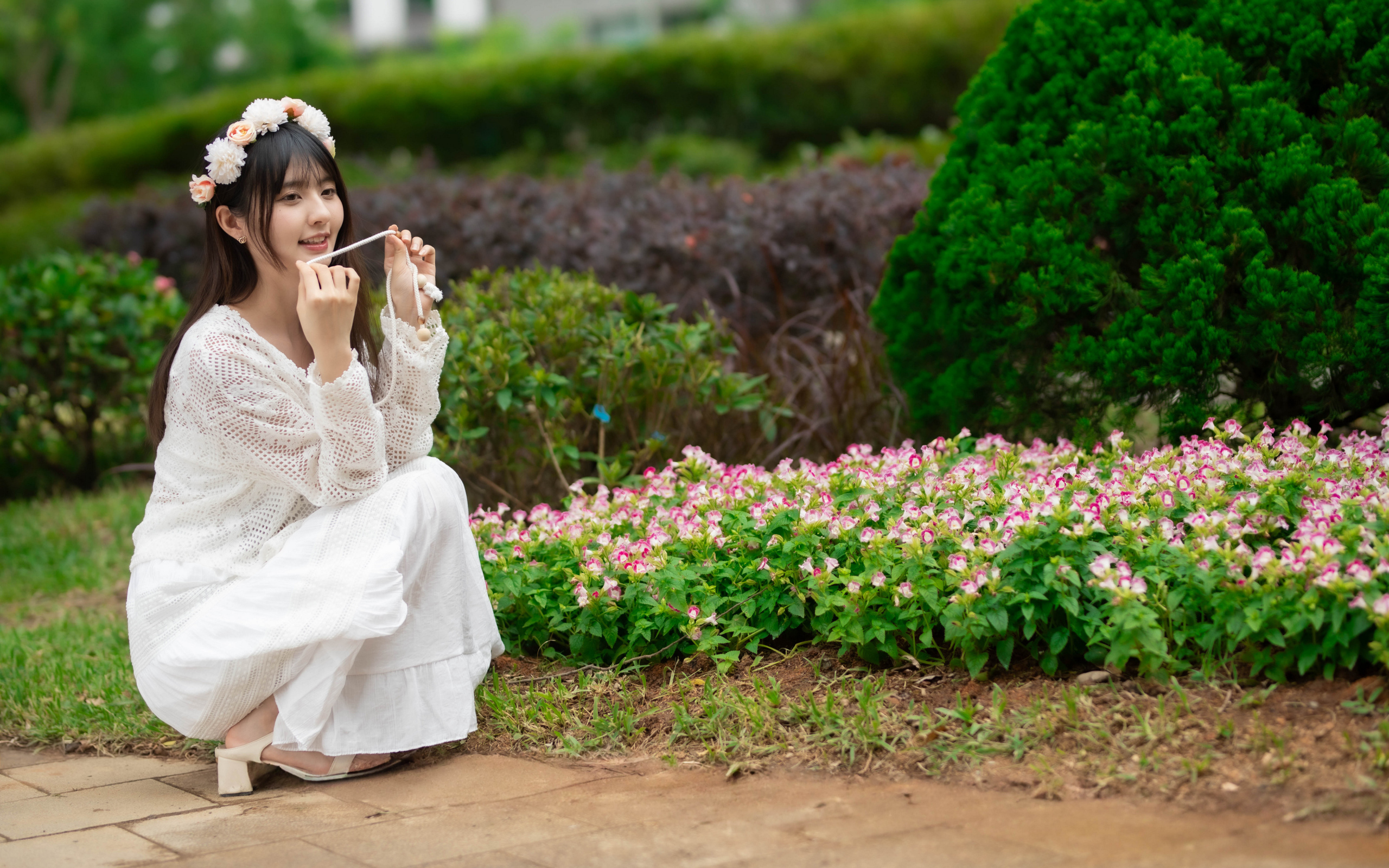 Милая азиатка в белом наряде сидит у клумбы с цветами