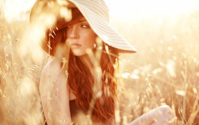 Рыжая девушка в шляпе сидит среди травы на поле