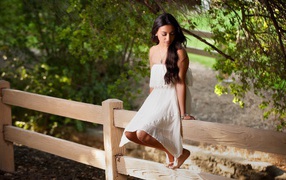 Брюнетка в белом платье сидит на заборе