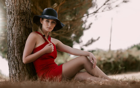 Красивая девушка в красном платье и черной шляпе сидит под деревом