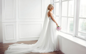 Beautiful girl in a white wedding dress with a bouquet is standing by the window