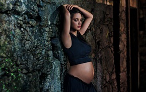 Беременная девушка в черном наряде у стены 