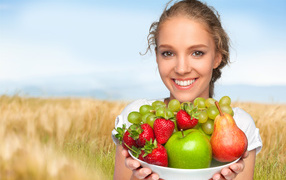 Smiling young lady with a plate with apples, grapes, strawberries and a pear in hands