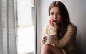 Молодая девушка в свитере сидит с чашкой на подоконнике