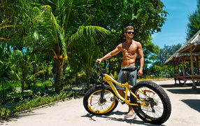 Красивый мужчина в очках с велосипедом у зеленых пальм