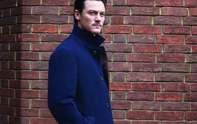 Актер Люк Эванс в синем плаще у кирпичной стены