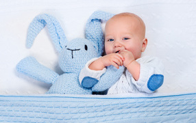 A cute baby lies in a crib with a blue knitted hare