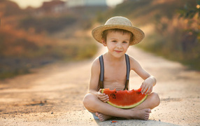 A little boy in a hat sits on the road with a slice of watermelon