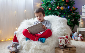 A little boy is sitting in a soft armchair by the New Year tree