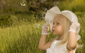 Маленькая девочка в большой белой шляпе пускает мыльные пузыри
