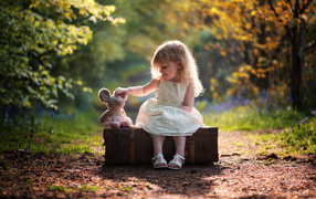 A little girl in a white dress is sitting on a big suitcase with a toy