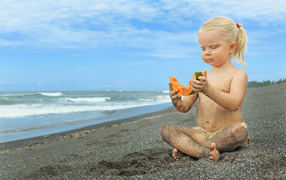 Маленькая девочка сидит на песке у моря с кусочком дыни