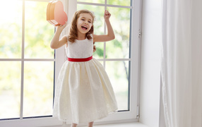 Маленькая девочка с коробкой в форме сердца в руках стоит на подоконнике