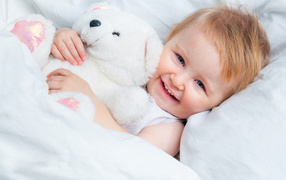 Улыбающийся ребенок лежит в кровати с белым плюшевым медвежонком