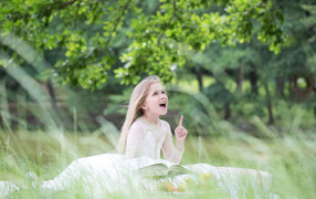 Красивая маленькая девочка в белом платье с книгой сидит на траве