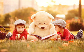 Big teddy bear with book and little boys lie on green grass