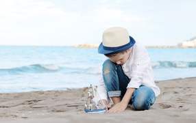 Маленький мальчик в шляпе играет с игрушечным корабликом на песке