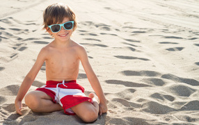 Little boy with glasses sits on the sand on the beach