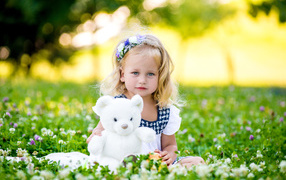 Маленькая милая девочка с игрушкой сидит на зеленой траве с цветами