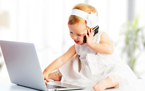 Маленькая девочка в белом платье сидит с телефоном у ноутбука