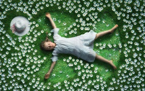 Маленькая девочка в белом платье лежит на зеленой траве с ромашками