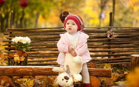 Маленькая девочка с плюшевым мишкой сидит на деревянной лавке осенью