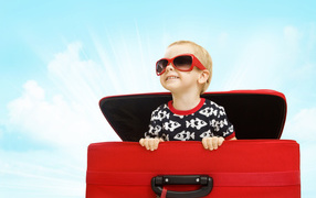 Маленький улыбающийся мальчик в очках сидит в красном чемодане