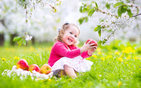 Маленькая улыбающаяся девочка сидит на зеленой траве с яблоками