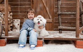 Маленькая улыбающаяся девочка сидит с белым пуделем на крыльце дома зимой