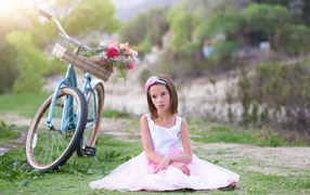Грустная девочка в красивом платье сидит на траве с велосипедом