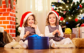 Две улыбающиеся девочки близняшки с новогодним подарком под елкой