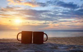 Две чашки стоят на песке на фоне рассвета 