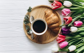 A cup of coffee with a croissant on a table with a bouquet of tulips