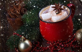 A cup of hot cocoa with marshmallow pieces and a bayanne on a table with a fir branch