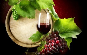 A glass of wine on the table with a bunch of grapes and a barrel