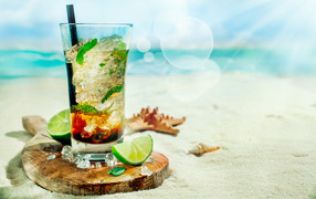 Cocktail in a glass with lime and ice cubes stands on the sand on the beach