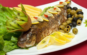 Baked fish with olives, lemon, kiwi, corn and lettuce on a white plate