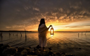 Девушка в белом платье стоит у воды с фонарем