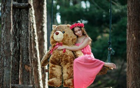 Красивая девушка азиатка в розовом платье сидит на качели с большим игрушечным медведем