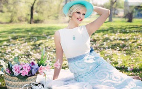 Красивая блондинка в голубой шляпке сидит в парке на траве