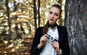 Красивая девушка с фотоаппаратом в руках стоит у дерева