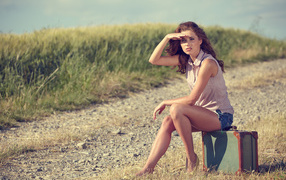 Красивая молодая девушка сидит на чемодане на дороге