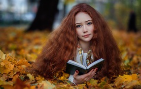 Рыжеволосая девушка с книгой лежит на опавших листьях осенью 